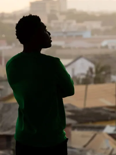 Silhouette of a person in a green shirt with arms crossed looking out over a densely built, hazy cityscape.